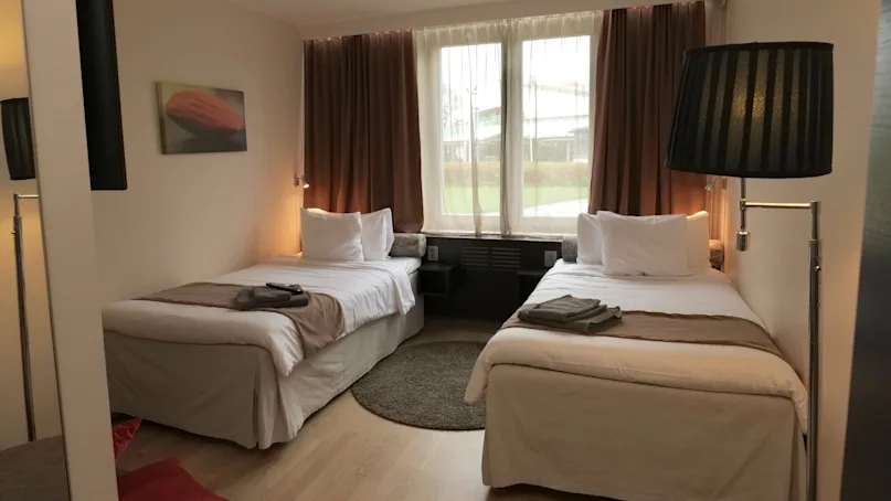 Twin beds are neatly arranged under a large window with drawn brown curtains, in a cozy hotel room featuring wooden flooring and a wall-mounted painting.
