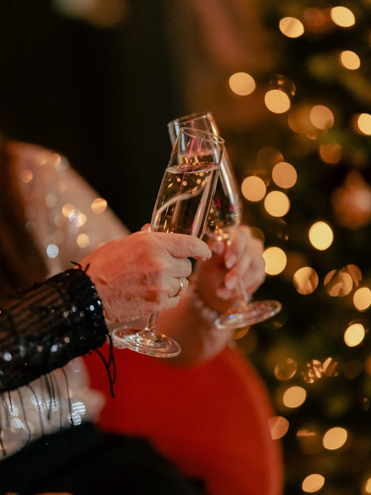 Two hands clinking champagne glasses, celebrating in a cozy setting with a blurred, illuminated Christmas tree in the background.