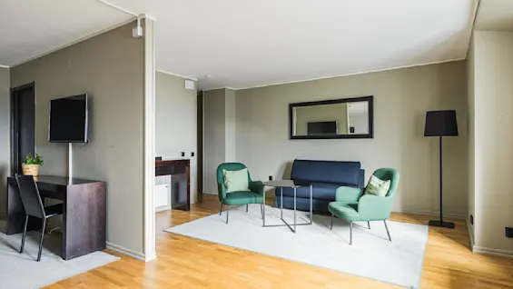 En moderne stue med grå vegger har en koselig sittegruppe med blå sofa og grønne stoler på et lyst teppe. Et speil over sofaen reflekterer lyset fra store vinduer.