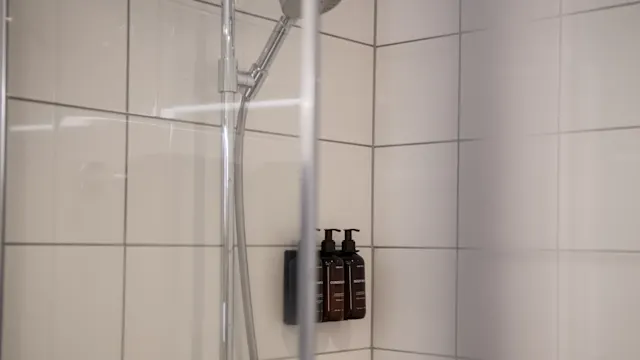 A showerhead is attached to a tiled wall, with three mounted pump bottles labeled "Shampoo," "Conditioner," and "Body Wash," set within a glass shower enclosure.