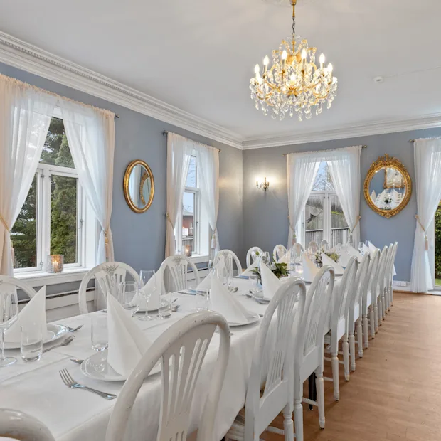 Et vakkert dekket langbord med hvite stoler i et lyst rom med lyseblå vegger, store vinduer og en glitrende lysekrone.