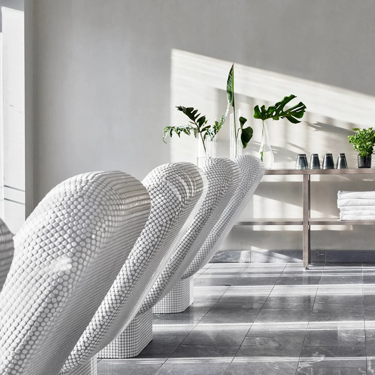 White textured seats line up in a modern, sunlit space with green plants and towels on a shelf.