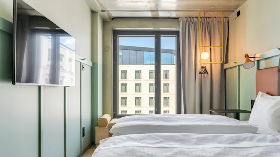 Et hotelværelse har to pænt redte senge med hvidt sengetøj ved siden af et stort vindue. En moderne lampe hænger fra en grøn væg, der også har et monteret TV.