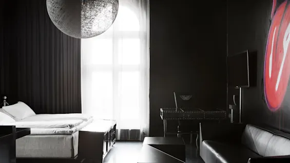 A modern bedroom with a large spherical light fixture hangs over a neatly made bed. To the right, there's a sofa, desk, and TV. The room is dimly lit with dark decor.