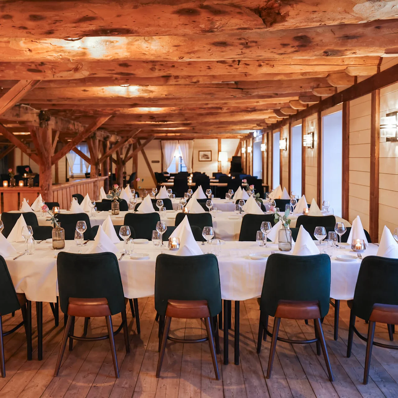 Dining tables set with white tablecloths and wine glasses; nestled in a rustic room with wooden beams and warm lighting, creating an inviting atmosphere.
