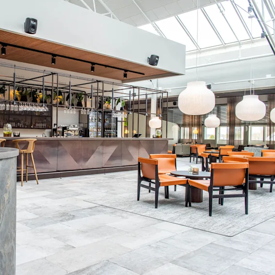 Bardisk med hängande vinglas och stolar, omgiven av trädekorationer och växter. Ljust område med modern belysning, orange stolar och bord i en öppen, rymlig miljö.