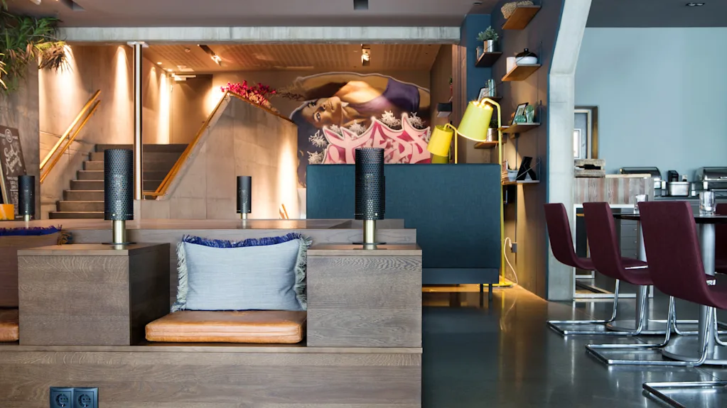 Ett modernt restauranginteriör med träbänkar och läderkuddar i förgrunden; dekorlampor och hyllor pryder väggarna, medan trappor leder till en mural som visar en liggande figur.