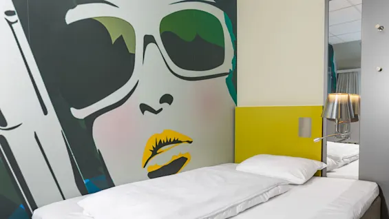 A bed with white linens is positioned against a colorful mural depicting a face with sunglasses and yellow lips. A metal lamp attached to a mirrored wall is nearby.
