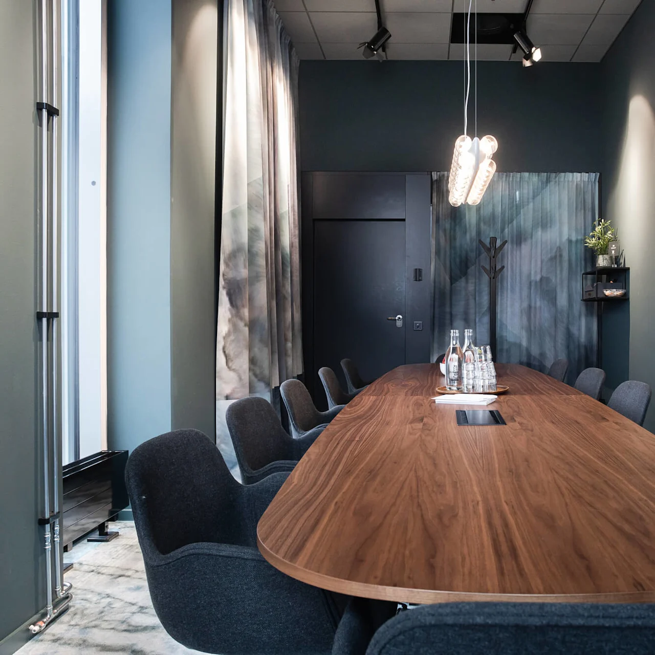Ett långt konferensbord av trä, omgivet av grå stolar, håller vattenflaskor och glas under hängande lampor. Rummet har mörka väggar, gardiner, en kapphylla och väggskåp.