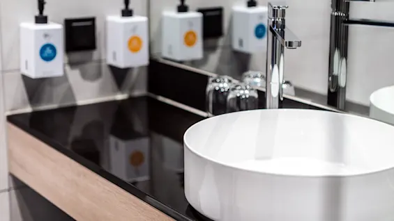En vit keramisk handfat ligger på en svart bänkskiva och reflekterar kromkran och två tvåldispensrar på väggen. Glas står bredvid handfatet i ett modernt badrum.