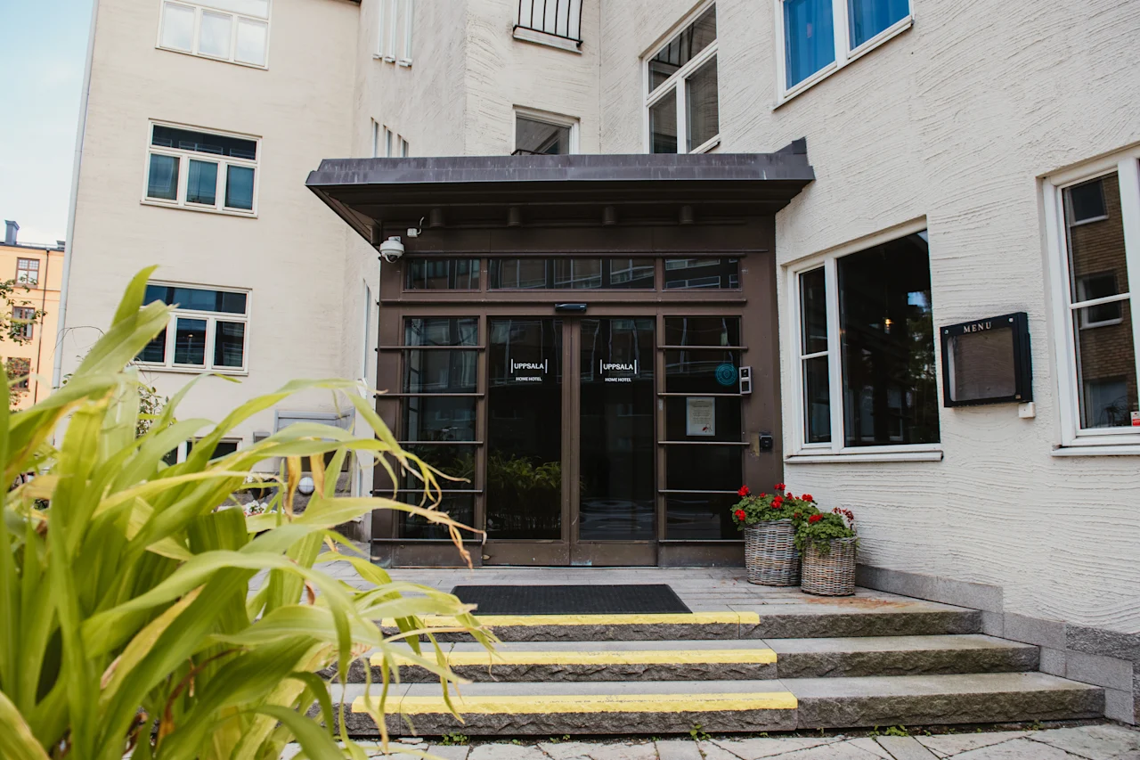 Entrance to Home Hotel Uppsala with glass doors, steps, and potted flowers. A menu sign is visible on the wall.