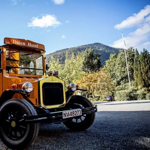 An antique yellow bus with "Dalen Hotel" sign and license plate NV48273 is parked on a gravel driveway, surrounded by a yellow building and lush greenery under a blue sky.