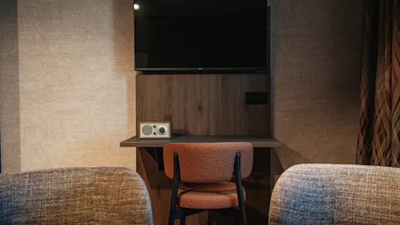 A hotel room corner with a wall-mounted TV above a desk, a chair, and two armchairs, ready for use.