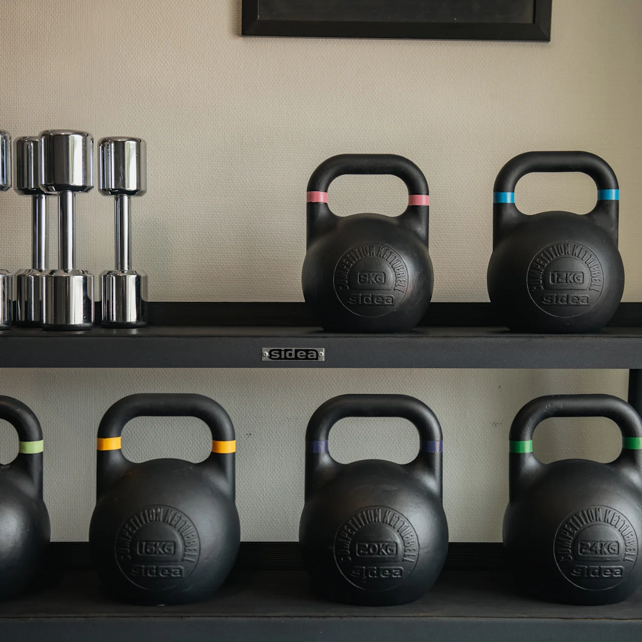 A set of kettlebells with colored bands and dumbbells sit neatly on a black shelf in a gym-like environment.
