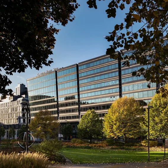 En modern glasbyggnad med skylten "Clarion Hotel Sign" står omgiven av frodiga gröna träd och en grästäckt park, med en person som promenerar längs en gångväg.