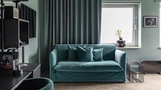 Et koselig rom med en turkis sofa, mørke gardiner og et skrivebordsområde, som skaper et avslappende og funksjonelt rom.