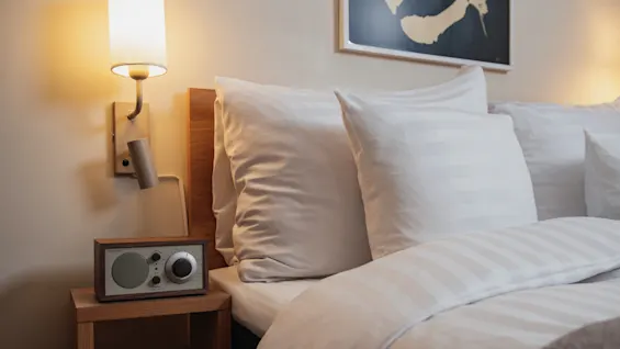 Bed with white pillows and duvet, beside a wooden nightstand holding a radio. Wall-mounted lamp illuminates the scene, and a framed artwork hangs above, creating a cozy bedroom atmosphere.