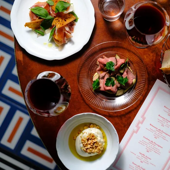A table with plates of assorted Italian dishes like sliced meat, bruschetta, and burrata; surrounded by glasses of red wine and a menu titled "SECOND." Features a tiled floor design.