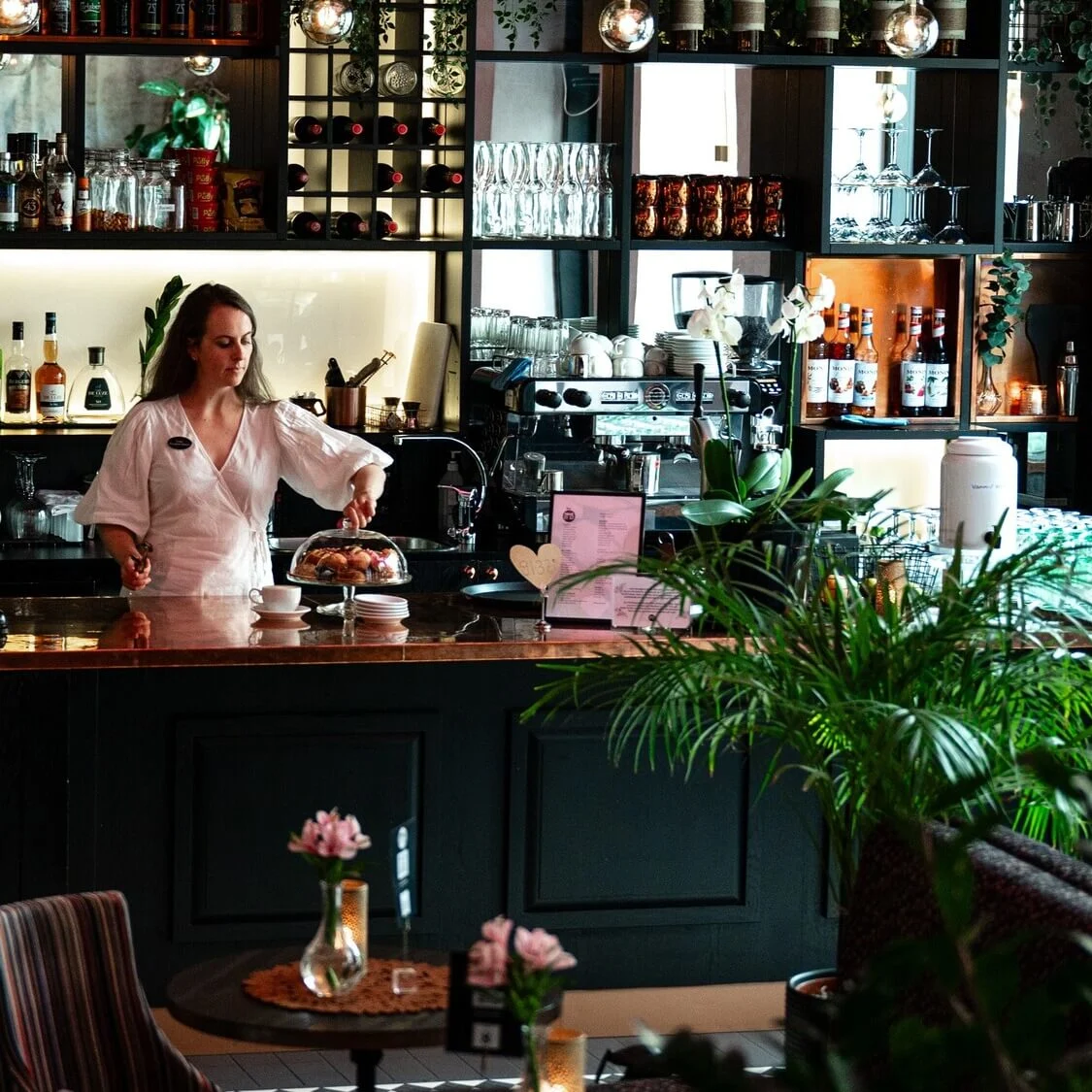 En bartender arrangerer kaker under en glassklokke i en koselig bar. Flasker og glass står pent på rad på hyllene bak henne, omgitt av frodige planter og myk belysning.