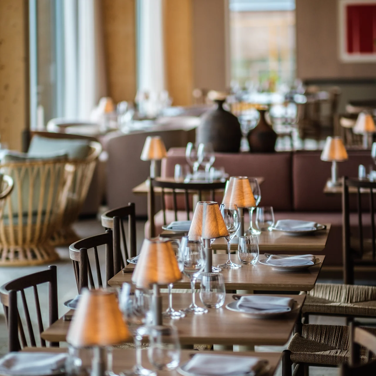 Tables are set with plates, glasses, and small lamps, creating an elegant atmosphere in a warmly lit restaurant with wooden furniture and large windows.