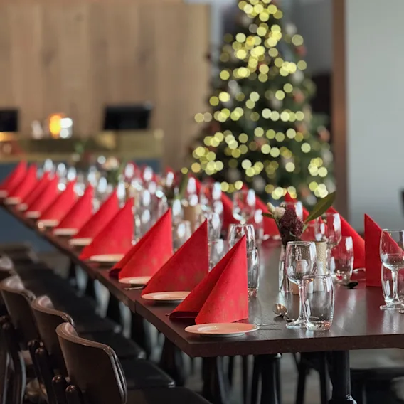Et langt bord dekket for et festmåltid med røde servietter og glass, med et uskarpt juletre i bakgrunnen.
