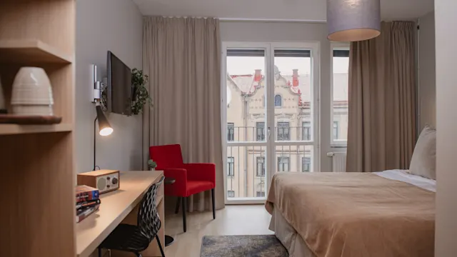 Ett modernt hotellrum med dubbelsäng, skrivbord och stol. Ett fönster med beige gardiner visar utsikt över utsmyckade byggnader utanför. Varm belysning och inredning skapar en mysig atmosfär.