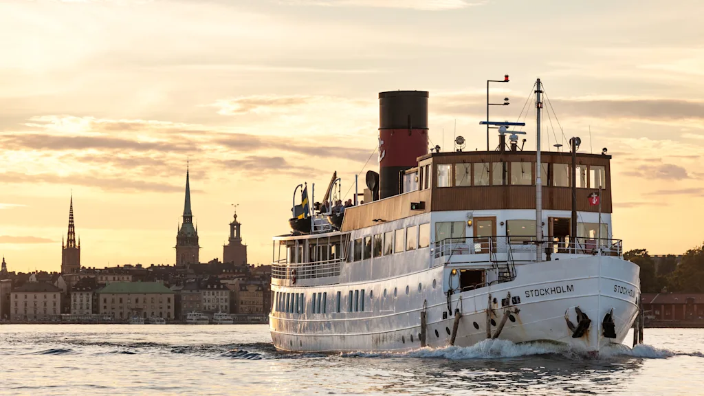 A Stockholm archipelago boat, named STOCKHOLM, sails on the water with a city skyline under a warm, glowing sky.