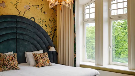 A bed is placed against a floral wallpaper and large arched headboard, adorned with decorative pillows. A chandelier hangs above, and a bay window offers a view of greenery outside.