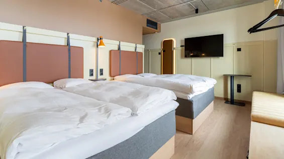 Tre fuldt opredte senge med hvide lagner står i et moderne hotelværelse med betonloft, trægulv, vægmonteret tv og minimalistisk indretning.