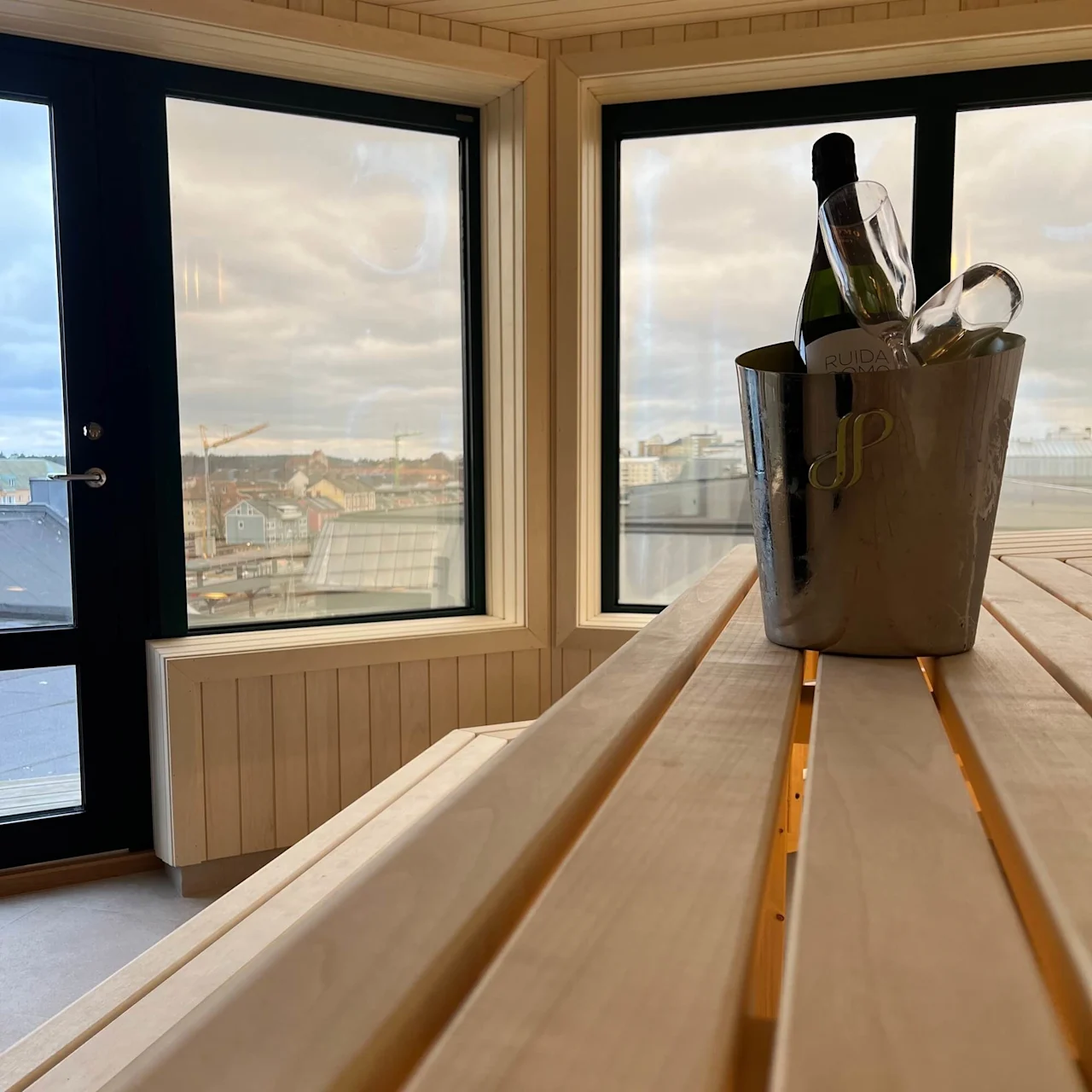 A champagne bottle in an ice bucket on a wooden table, against a backdrop of windows showing an urban skyline.