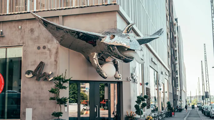 En stor, skinnende mantaskulptur stikker ut fra en bygningfasade over en døråpning, med kafébord i nærheten på en bygate. Tekst: "Ax" og "AVEQIA".