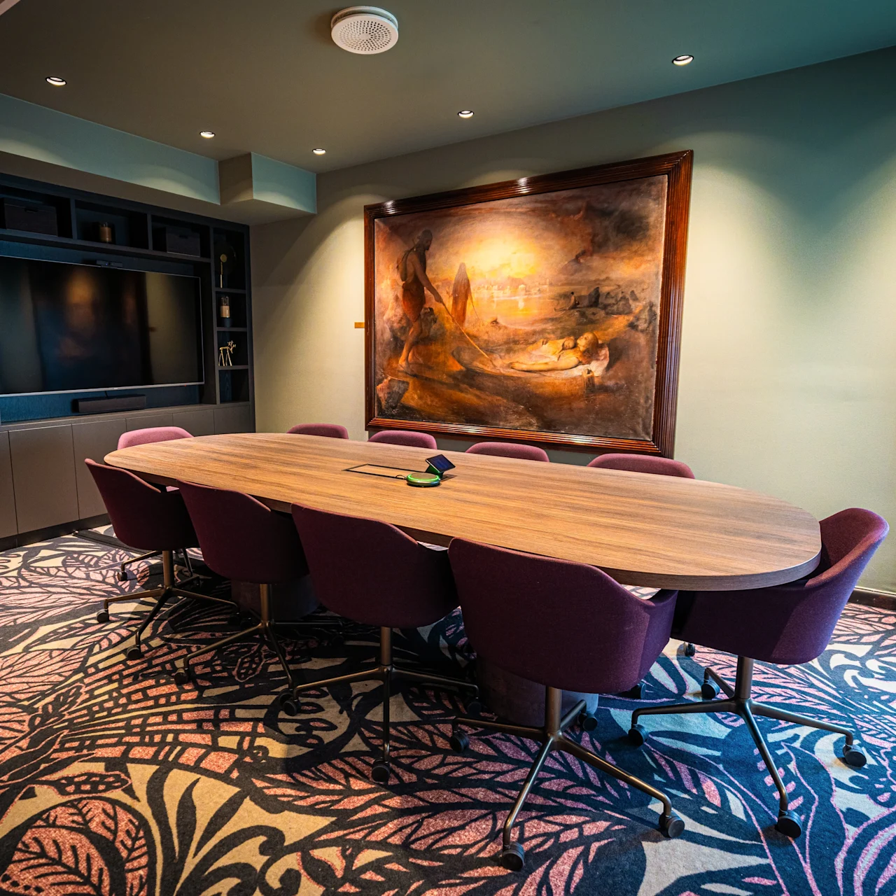 A large wooden conference table with purple chairs is placed in a room with intricate patterned carpet. A large painting hangs on the green wall, and a television is mounted nearby.