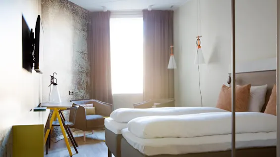 A modern hotel room features a neatly made double bed, a wall-mounted TV, and a desk with a lamp. Natural light filters through partially drawn curtains.