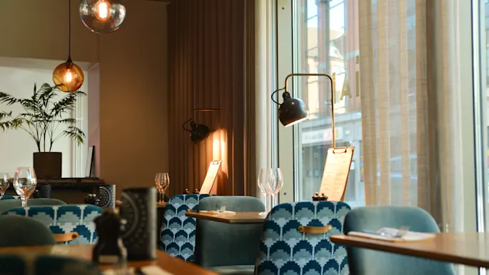Elegant restaurant with blue patterned chairs, illuminated by warm pendant lamps, near large windows. Tables are set with wine glasses and menus under natural and ambient lighting.