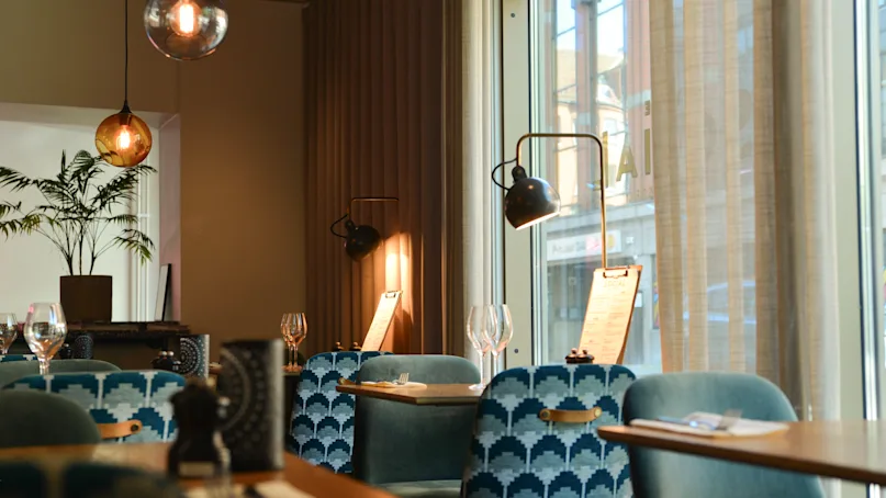 Elegant restaurant with blue patterned chairs, illuminated by warm pendant lamps, near large windows. Tables are set with wine glasses and menus under natural and ambient lighting.