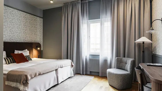 Ett väl inrett sovrum med två sängar, eleganta gardiner, matchande fåtölj, skrivbord och belysta lampor som förmedlar en bekväm och stilfull interiör.