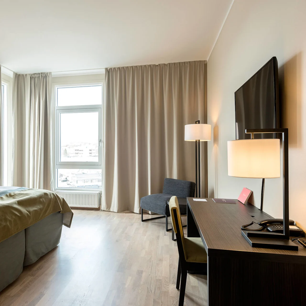 Siististi järjestetty hotellihuone, pedattu sänky, työpöytä tuoleineen, läppäri ja puhelin, sekä ikkuna kaupunkinäkymällä.