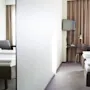 Two neatly made beds are on opposite sides of a light-filled hotel room, separated by a partial wall. A desk with a lamp, chair, and TV is visible.