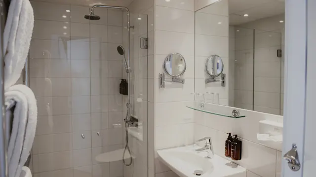 Ett vitt kaklat badrum med regndusch, modern handfat, stor spegel, två runda väggspeglar och prydligt vikta handdukar till vänster.