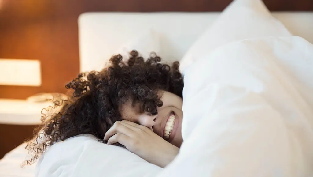 Happy woman taking it easy in a lush hotel bed, knowing she won't be downgraded in all of 2021.