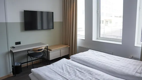 En veggmontert TV vender mot to pent oppredde enkeltsenger i et lyst rom. Ved siden av står et enkelt skrivebord med en stol under og et stort vindu ved siden av.