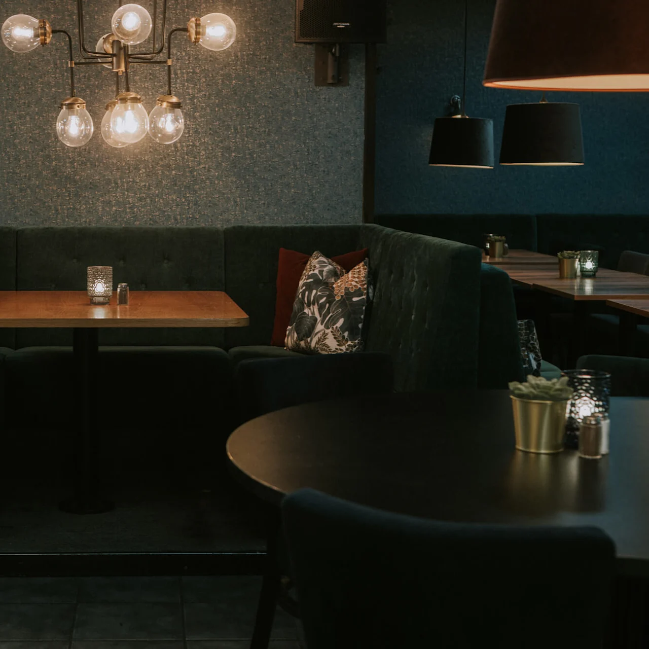 Et hyggeligt restauranthjørne med plys siddepladser og borde, svagt belyst af stilfulde loftslamper og små bordlys.