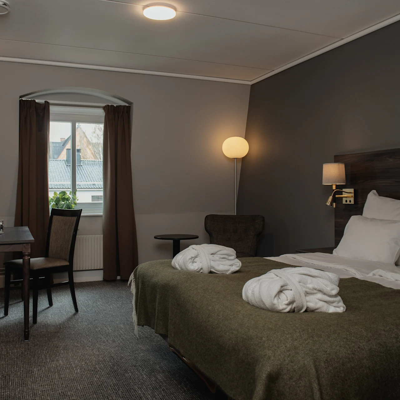 A bed adorned with white robes rests in a warmly lit hotel room, featuring a desk, chair, window with curtains, and a floor lamp in a cozy atmosphere.