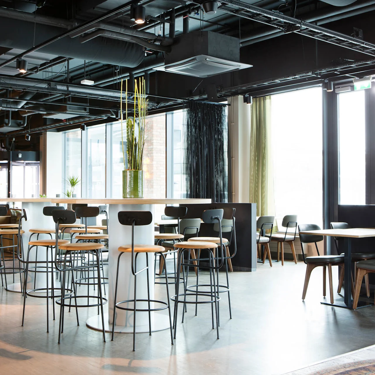 Modernt caféinteriör med bord och stolar, stora fönster som ger naturligt ljus. Industriellt tak och mjuka naturliga toner skapar en avslappnad atmosfär.