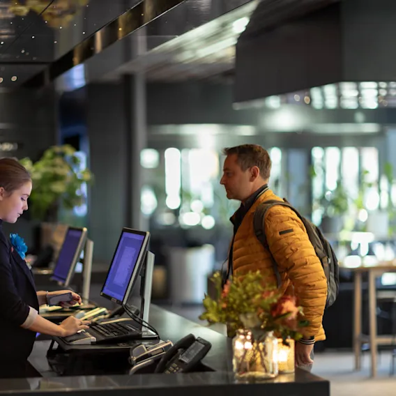 En receptionist använder en dator för att hjälpa en stående man med ryggsäck i en modern, välbelyst lobby dekorerad med växter och blommor.