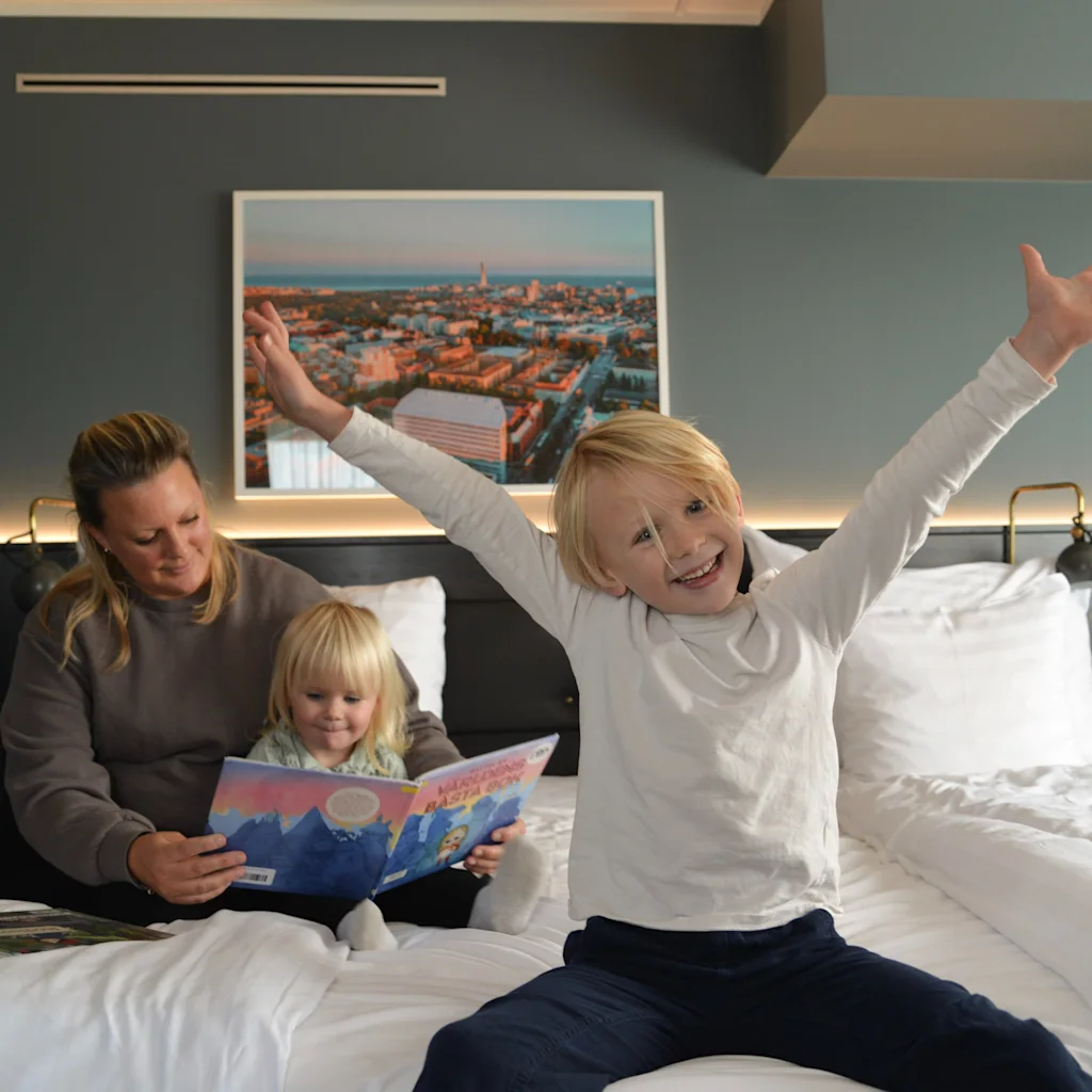 En familj njuter av en mysig stund i ett hotellrum, med en vuxen som läser en bok för ett barn och ett annat barn som glatt leker.