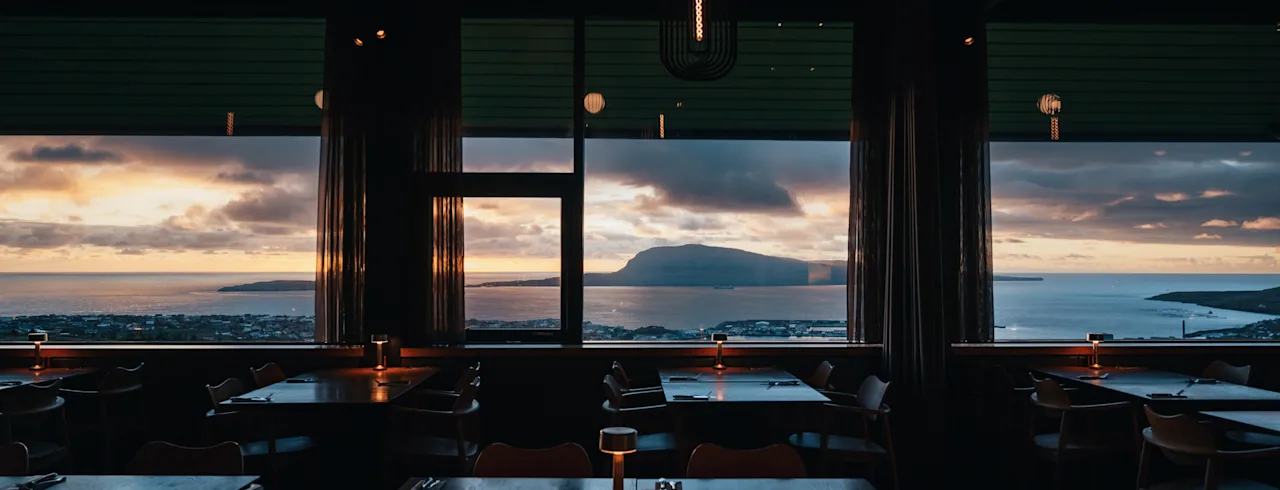 Restaurantinteriør med dekket bord og fantastisk utsikt over havet og øy ved solnedgang. Restauranter i Tórshavn.