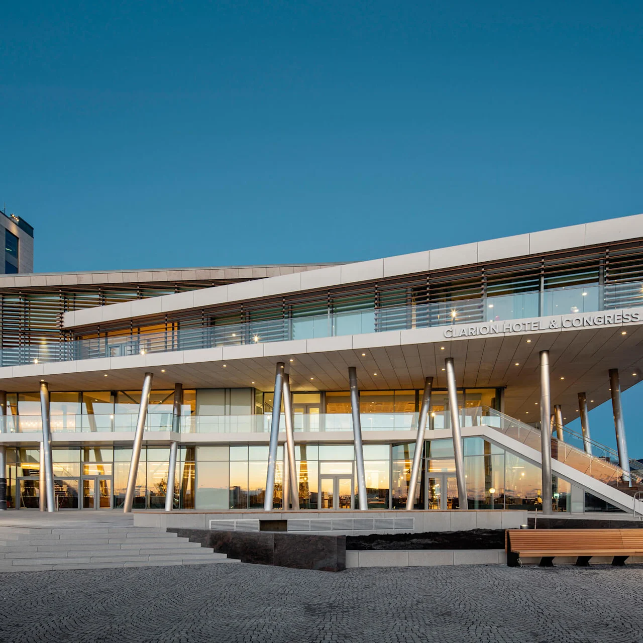 En moderne bygning med skrånende tak og glassvegger, støttet av sølvkolonner, står mot en skumringshimmel. Tekst: "CLARION HOTEL & CONGRESS SeaU".
