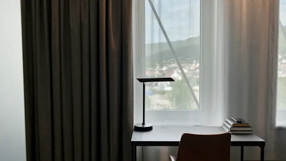 En koselig arbeidsplass med et skrivebord, lampe og bøker ved et vindu, med utsikt over en by og åser.