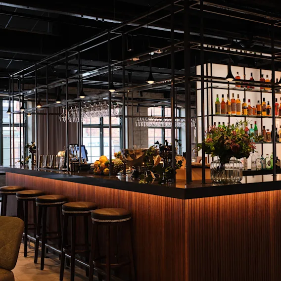 Barbänk med höga pallar som visar glas och flaskor; en vas med blommor lägger till dekoration. Baren finns i en modern, industriell-chic miljö med stora fönster.